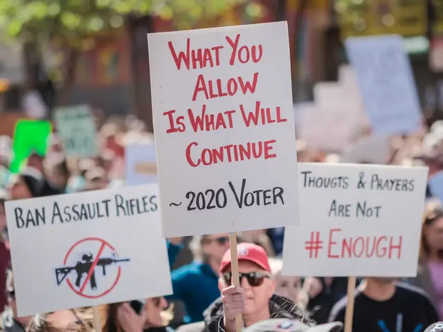 The image shows a crowd of people holding signs in front of a building, with trees in the...