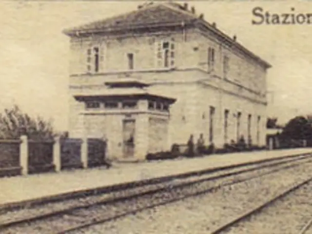 The image shows an old photo of a train station with a building in the center, surrounded by trees...