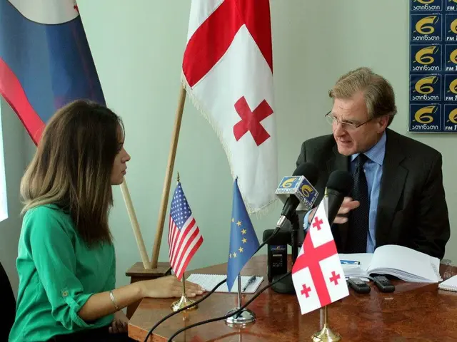 The image shows a man and woman sitting at a table with flags in front of them. The man is wearing...