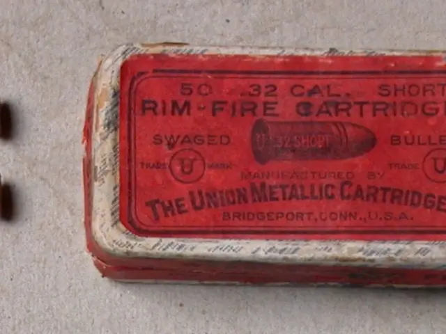 The image shows a red box of ammunition sitting on top of a table, with two bullets beside it. The...