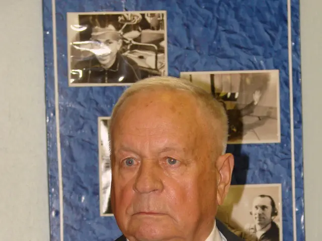 The image shows a man in a suit and tie standing in front of a wall adorned with photos. He has a...