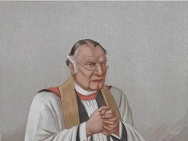 The image shows a man in a priest's robes standing at a podium with a book in front of him. He has...