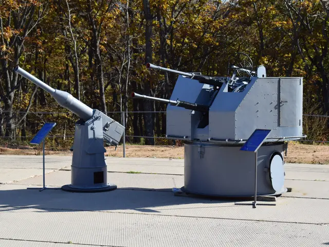 The image shows a large machine gun sitting on top of a parking lot surrounded by trees and a fence...