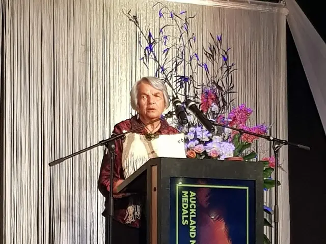 The image shows a woman standing at a podium in front of a microphone, surrounded by a bouquet of...