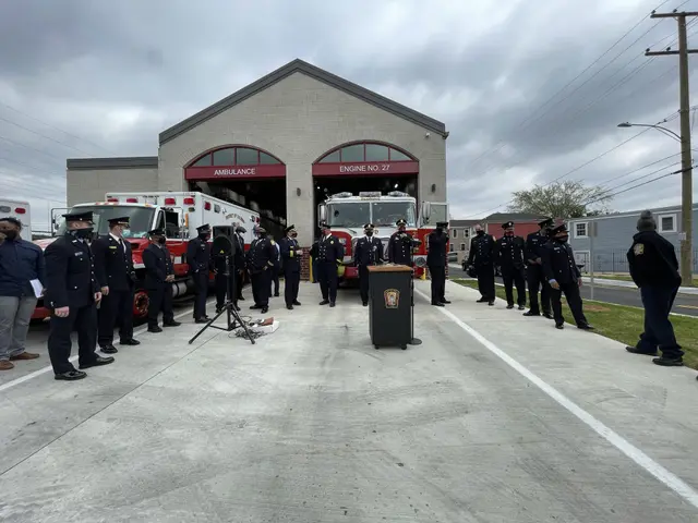 The image shows a group of people standing in front of a fire truck, with a podium and microphone...