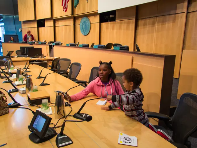 The image shows two children sitting at a long table in front of a microphone, surrounded by...