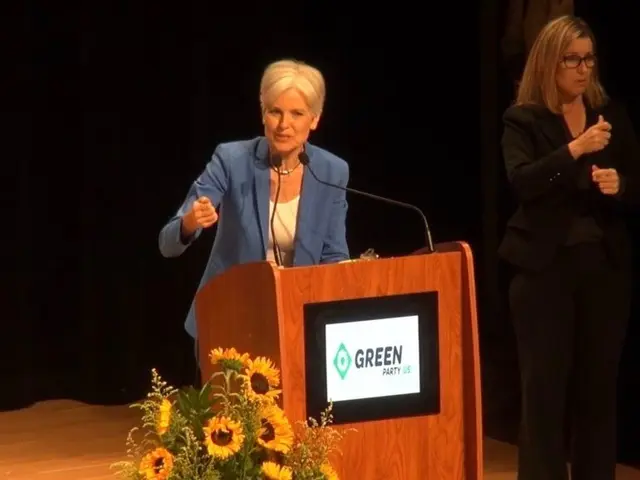 The image shows two women standing at a podium in front of a microphone, with a screen attached to...