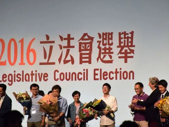 The image shows a group of people standing on top of a stage, holding flower bouquets in their...
