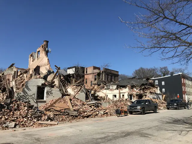 The image shows a collapsed building on the corner of a street, with vehicles on the road, people...