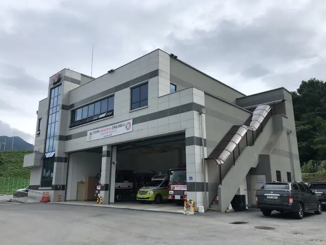 The image shows a fire station with cars parked in front of it. The building has windows and a...
