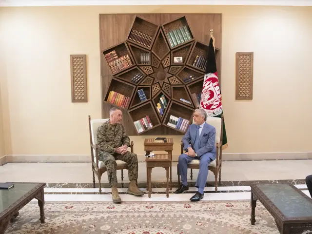 The image shows two men sitting in chairs talking to each other in a room. On the tables in front...