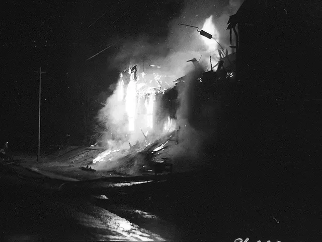The image shows a black and white photo of a fire burning in the middle of a street, with street...