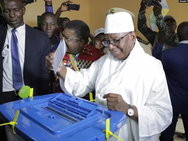 The image shows a man in a white suit and hat putting his vote into a blue ballot box surrounded by...