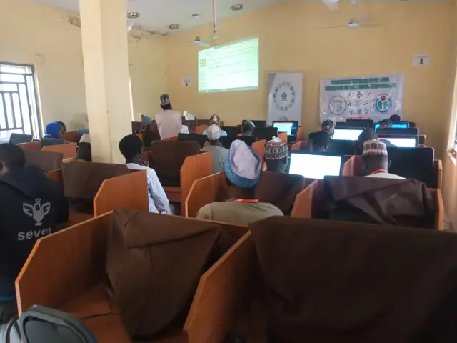 The image shows a group of people sitting in chairs in front of computers in a room with a wall in...