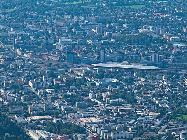 The image shows an aerial view of a city with lots of buildings, trees, roads, and grass. The city...