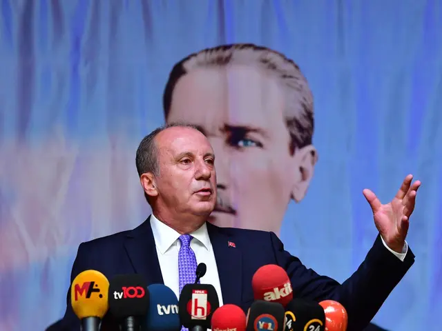 The image shows a man in a suit and tie standing at a podium with microphones in front of him and a...