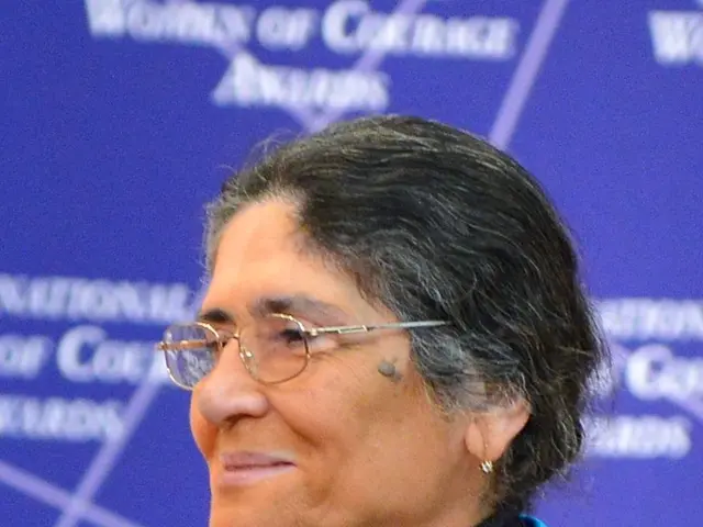 The image shows a woman wearing glasses and a colorful jacket standing in front of a blue wall with...