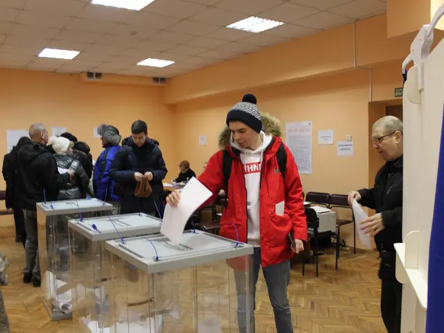 The image shows a group of people standing around a voting booth in a polling station. There are...
