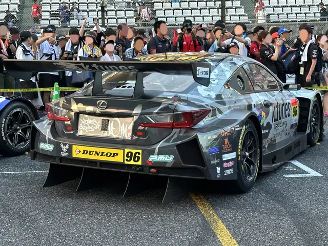 The image shows a Lexus RC F GT3 race car on the road surrounded by a group of people, some of whom...