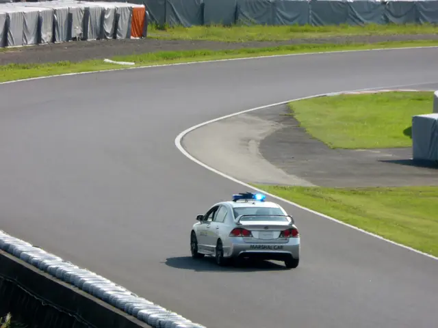 The image shows a police car driving down a race track, surrounded by grass on either side of the...