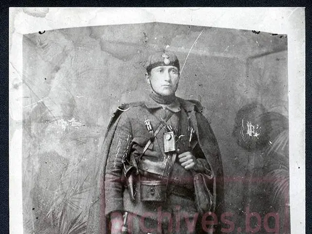 The image shows an old black and white photo of a man in uniform, wearing a cap and holding a...