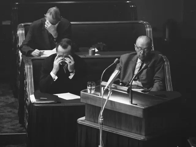 The image shows two men sitting at a podium in front of a microphone, surrounded by tables with...