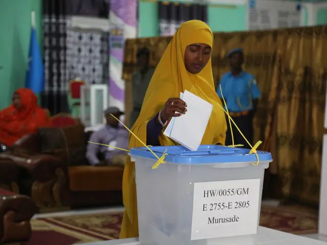 The image shows a woman in a yellow headscarf casting her vote at a polling station. She is...