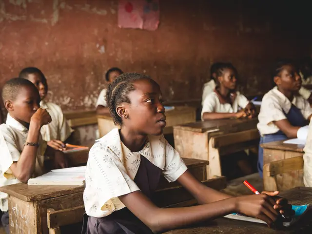 The image shows a group of children sitting at desks in a classroom, with books and pens on the...