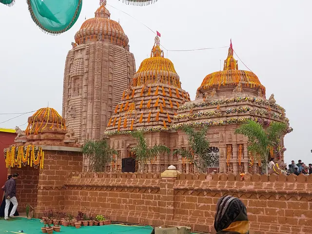 The image shows a group of people standing in front of a Hindu temple, surrounded by lush greenery...