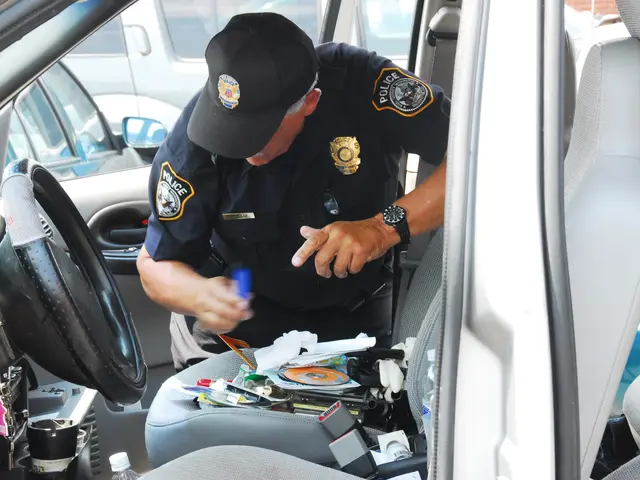 The image shows a police officer sitting in the driver's seat of a car. He is wearing a uniform and...