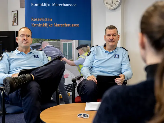 The image shows two police officers sitting at a table talking to a woman. On the table there are...