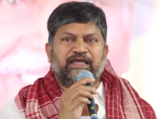 The image shows a man wearing a white shirt and a red shawl, speaking into a microphone in front of...