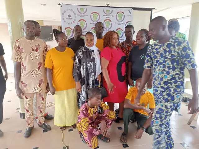 The image shows a group of people standing in front of a banner with text on it, surrounded by...