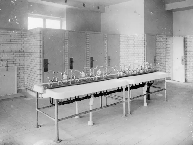 The image shows a black and white photo of a public restroom with a row of sinks, taps, walls,...
