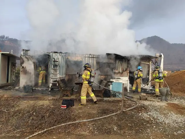 The image shows a group of firefighters wearing helmets and holding pipes, standing in front of a...