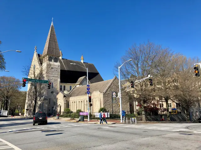 The image shows a large church with a steeple on the corner of a street, surrounded by buildings,...