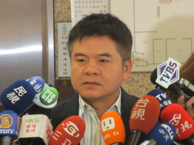 The image shows a man in a suit and tie speaking to reporters in front of a wall with a board...