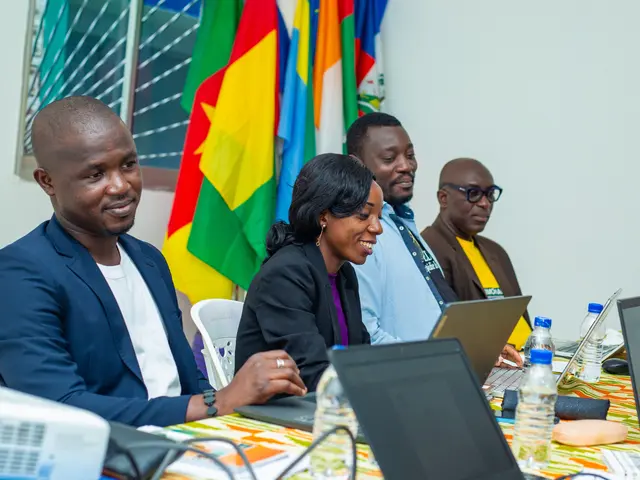 The image shows a group of people sitting around a table with laptops, bottles, mics, papers, and...