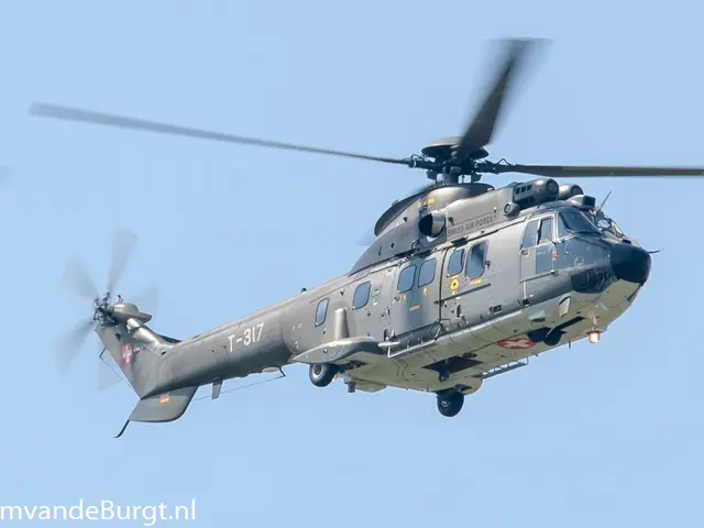 The image shows a military helicopter flying through a bright blue sky with its landing gear down,...