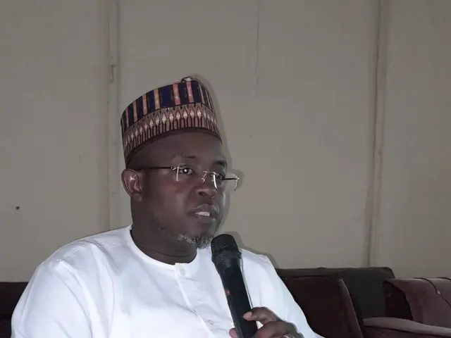 The image shows a man wearing a white dress and a cap on his head, sitting in a chair and speaking...
