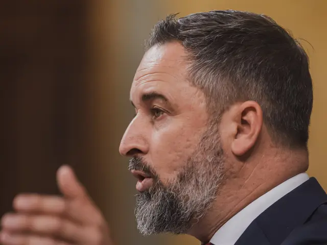 The image shows a man in a suit and tie speaking into a microphone in front of a blurred...