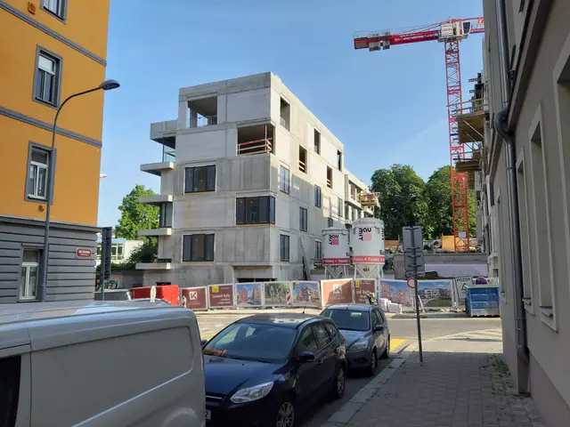 The image shows a street with cars parked on the side of it, buildings with windows, light poles,...