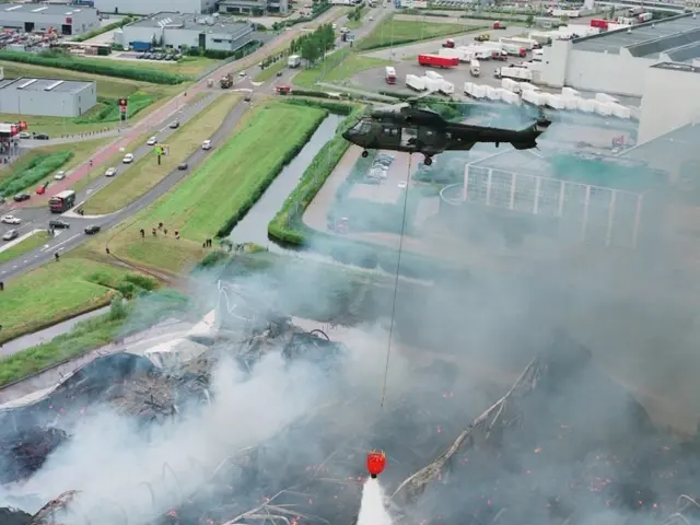 The image shows a helicopter flying over a large building that has been destroyed by fire, with...