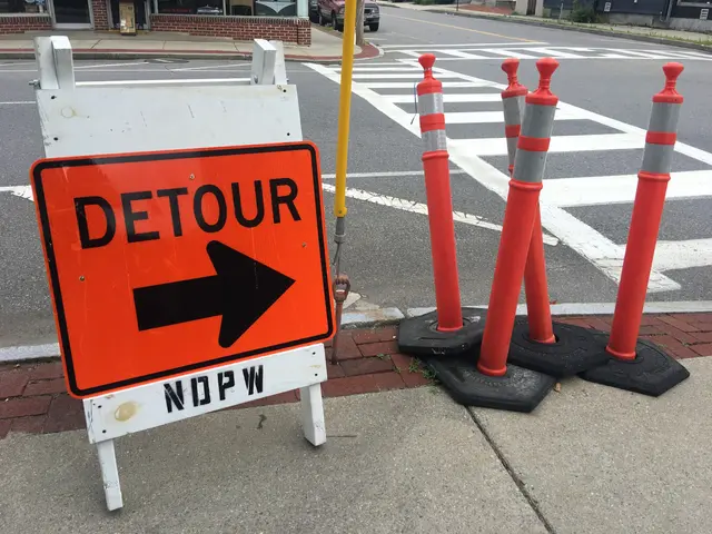 The image shows a detour sign sitting on the side of a street, surrounded by poles, vehicles,...