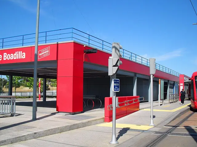 The image shows a red train traveling down tracks next to a red building, which appears to be a bus...