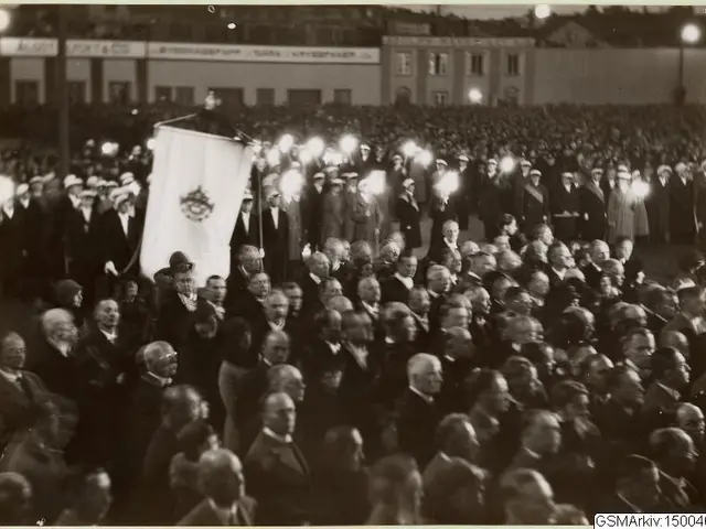 The image shows a large crowd of people standing in front of a building, with a person holding a...