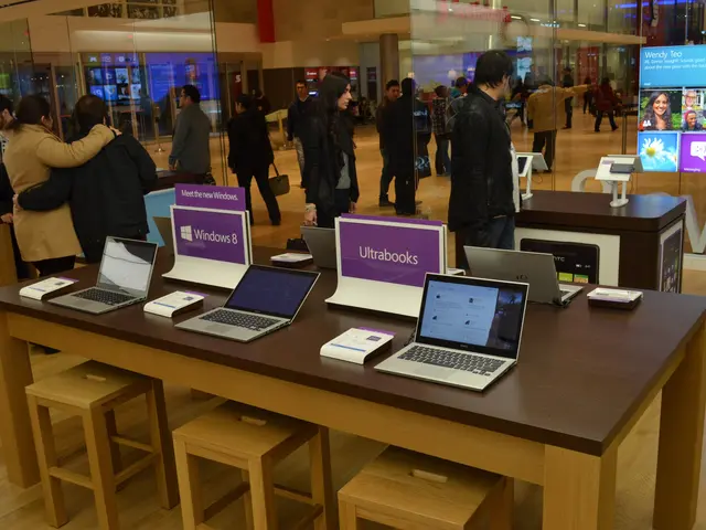 The image shows a group of people standing around a table with laptops on it, surrounded by stools,...