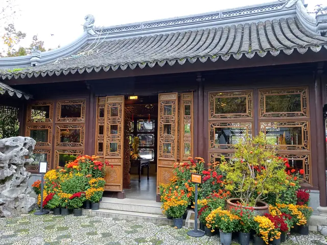 The image shows a Chinese building with a lot of flowers in front of it, surrounded by trees and a...