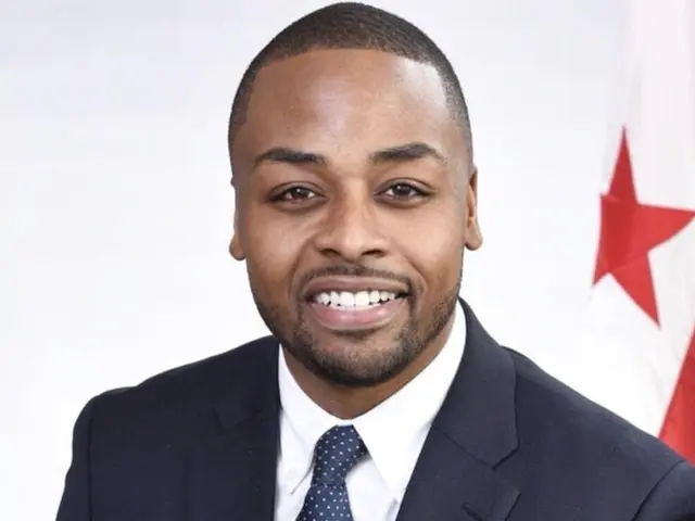 The image shows a man in a suit and tie smiling at the camera, with a white background and a red...
