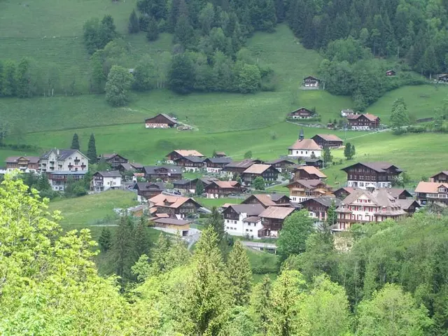 The image shows a small village nestled in the middle of a lush green valley, surrounded by trees...